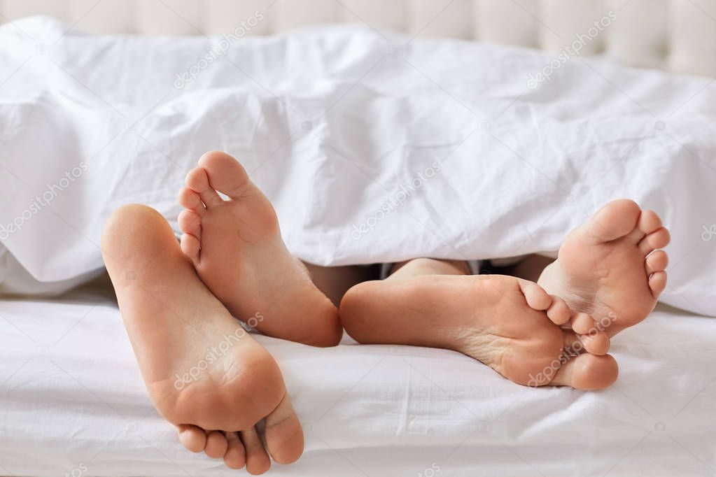 Female`s and male`s bare feet have rest under white blanket, recreat together, feels relaxation. Unrecognizable family couple sleep in comfortable bed. Bed time and good rest concept