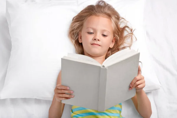 漂亮的小学龄前儿童读着有趣的故事 拿着翻开的书 躺在床上 Enjoyes 看书之前睡觉 有好的休息 聪明的小女孩读童话故事 儿童概念 — 图库照片