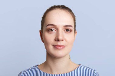 Yakın memnun güzel kadın portresi vardır saf deri, koyu renk gözleri, at kuyruğu bağlı saç, mavi arka plan üzerinde izole kamera, içine güvenle görünüyor. Güzel ve genç birbayan pozlar kapalı