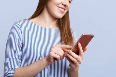 Cropped shot of young female with cheerful expression holds modern cell phone, messages with friends, connected to wireless internet, being always in touch, isolated over blue studio background clipart