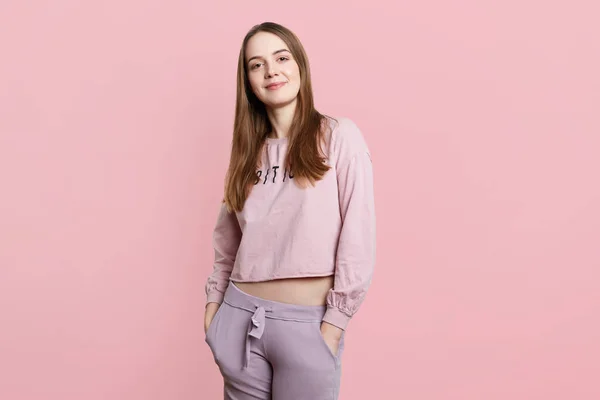 Slim sporty female model wears casual sport clothes, keeps hands in pockets, looks directly into camera, isolated over pink background. Fashionable young woman poses in studio, rests at home