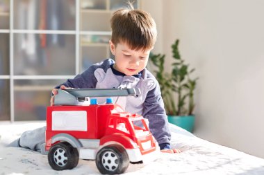 Portrait of small male with attractive look, plays with red toy automobile, sits on bed, spends free time in bedroom against cozy interior, likes cars, being future driver. Children and leisure clipart