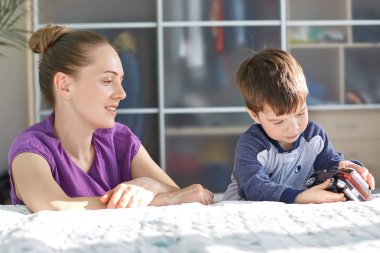 Ve çocuk kavramı gündeme getirme. Mor rahat t shirt, Genç ve güzel anne yatay atış onun küçük oğlu ilgilenir, yatak yatak odasında oyuncak araba ile birlikte oynamak, olumlu görünüme sahip