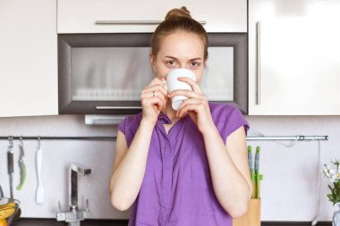 Kapalı atış hoş genç Sevimli Kadın giyer mor casual giyim, seyir, sabah, pozlar mutfak iç yalnız karşı saç topuz içecekler sıcak içecek vardır stong aromatik kahve sahiptir