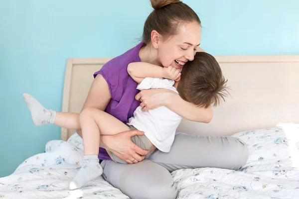 积极年轻有吸引力的母亲和她的小儿子玩耍 表达快乐 睡得开心 坐在柔软的床上 在家里享受平静的家庭气氛 母性概念 — 图库照片