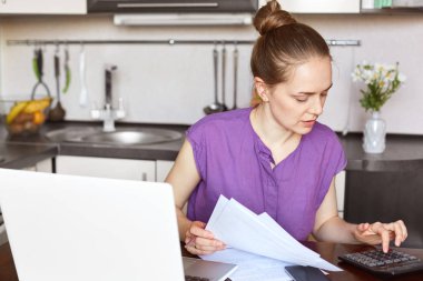 Meşgul çalışma kadın freelancer iş raporu yapar, rakamlar hesaplar ve belgeleri, dizüstü bilgisayar, kablosuz internet'e bağlı, eserlerinden oturur mutfak iç karşı tutar.
