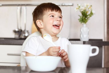 Çocuk ve beslenme kavramı. Mutlu küçük sevimli gülümseyen lezzetli mısır gevreği yiyor ve çay keyfi yerinde olmak, içecekler, mutlu annesine benziyor, mutfak oturur. Çok memnun küçük erkek çocuk