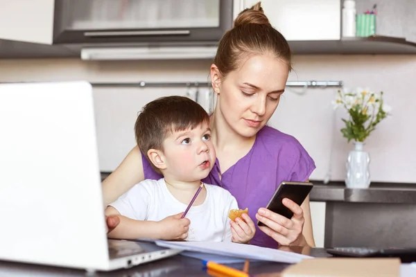 工作繁忙的母亲的照片试图解决财务问题 等待电话 持有智能手机 坐在孩子的工作场所 赚取钱的产假 周围的文件 距离工作 — 图库照片