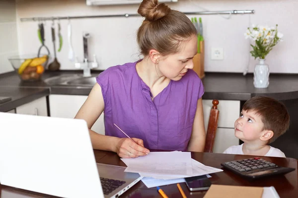 Horizontal shot of working mum being busy with documentation, makes financial report or calculates family budget, surrounded with papers and laptop, looks at her child and asks to be obedient