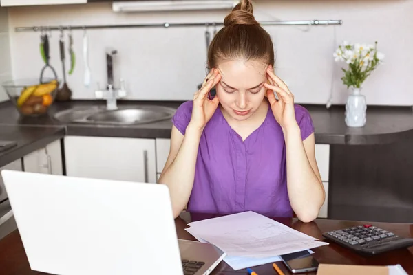 Overworked young female businesswoman has headache after sleepless night, works on business report, tries to calcualte figures, surrounded with calculator, laptop and documents, being at kitchen
