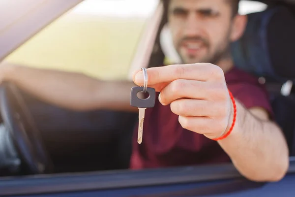 Handsome male holds car key while sits in automobile, sells his auto, travells in long journey. Man haulier drives on long distance. Focus on key. Horizontal shot. People and transportation concept