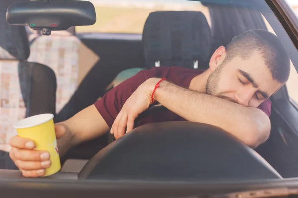 Fatigue overworked tired male driver has long trip in car, makes stop to have rest, has nap on helm, holds coffee in hands, looks sleepy. Long distance driver sleeps in automobile, has rest.