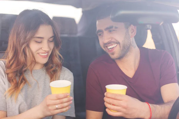 Glad female and male couple have coffee break in car, have long trip, enjoys beautiful views, spend vacation together, reach destination wih high speed. Family has trip or journey by vehicle