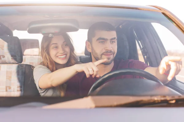 Excited female and male in car, points at windscreen, notice something funny on road, enjoy unforgettable trip in automobile, have good transport for travelling. People and reaching destination