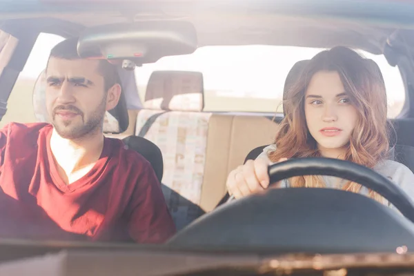 Couple in car on road trip: concentrated experienced female driver sits at wheel and her husband at front passanger seat, look in window with focused dreamy expressions. Transport, road and travel