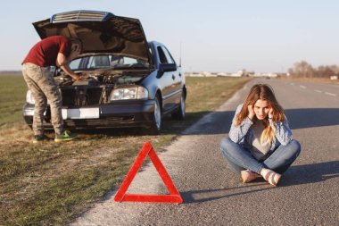 Breakdown alongside road. Male tries to repair car, call by cell phone in road assistance, can`t solve problem by himself, stressful woman sits crossed legs on asphalt near warning red sign. clipart