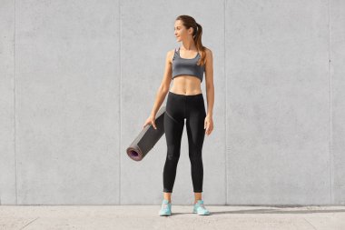 Tam vurdu uzunluğu ince kadın fitness beden, giyer üst, tozluk ve spor ayakkabıları ile egzersiz minderi, spor salonunda egzersiz olacak poz veriyor gri beton duvara tutar. düşünceli bir yana görünüyor
