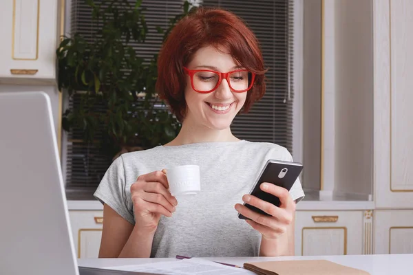 Cheerful fashionable woman with red hair, glad to view photos on social websites, has coffee break after working at laptop computer, wears stylish red spectacles and casual grey t shirt works remotely