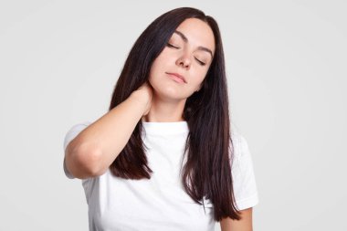 Fotoğraf yorgun esmer kadın hisseder ağrı boyun sedanter yaşam tarzı, olduğu gibi fiziksel aktivite ihtiyacı, gözlerini kapatır, uyku istiyor, casual Beyaz tişörtlü, standları stüdyo duvar karşı giymiş dinlen