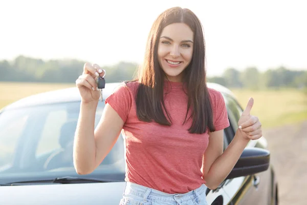 户外拍摄的满意美丽的黑发女车主的车 喜欢和高兴她的购买 持有钥匙和保持拇指提出 有愉快的表达 站在新汽车附近 要开车 — 图库照片