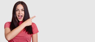 Pembe t gömlek giymiş siyah saçlı sürpriz esmer kadının yatay atış ile işaret parmağı asie, puan gösterir, promosyon için boş alan inanılmaz bir şey fark eder. Şuraya bak!