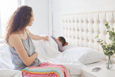 Pijama kıvırcık güzel kadın yatağın rahat pijama giymiş, tarafında oturur, kocası uyanık, relatioships ile ilgili sorunlar var, rahat yatak odası, Aile ve kavram uyku içinde teşkil etmeye çalışır