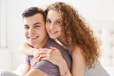 Olumlu ifade ile evli yeni evliler evde kapalı günler. Kıvırcık kadın kucaklayan aşk kocasıyla gülümseyen, rahat kıyafetler, hızlı mutluluk giyinmiş. İnsanlar ve ilişki kavramı