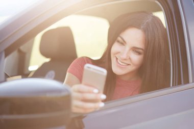 Neşeli iyi looing kadın arabada oturur, modern akıllı telefon tutar, selfie yapar, sosyal ağlar fotoğrafları paylaşır, ücretsiz internet bağlantısı ve yüksek hızlı sahiptir. Ulaşım ve insanlar kavramı