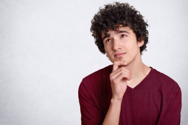 Ayrı tutma çene, derin düşünceler içinde varlık hakkında bir şey, contemplates düşünceli kıvırcık adam çalışır rahat t gömlek, beyaz beton duvara reklam için pozlar giymiş düşünülerek karar