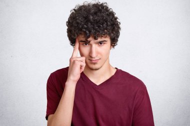 Headshot düşünceli ciddi yakışıklı erkek, siyah kıvırcık saçlı, işaret parmağı Tapınağı üzerinde tutar, rahat t gömlek giyer, beyaz beton duvara teşkil etmektedir. Genç hakkında bir şey contemplates