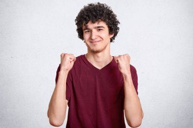 Yakışıklı çekici bakışla kapalı atış içinde yumruklarını kaldırdı ellerini tutar, hoş gülümseme ve mutlu ifade, kıvırcık siyah saçlı, beyaz beton duvardan izole gücünü güvenen vardır