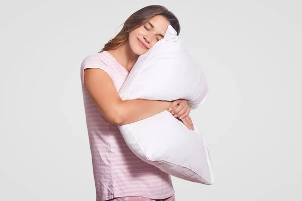 高兴的年轻欧洲妇女的照片在良好的心情 拥抱软枕头 穿着睡衣 构成反对白色背景 放松的青少年睡在室内 休息和生活方式概念 — 图库照片