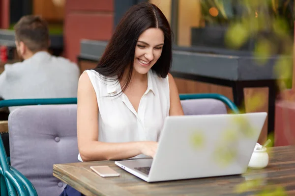 Positive female blogger with dark straight hair, works on laptop computer, connected to wireless internet in outdoor restaurant. Beautiful young woman chats in social networks with best friend