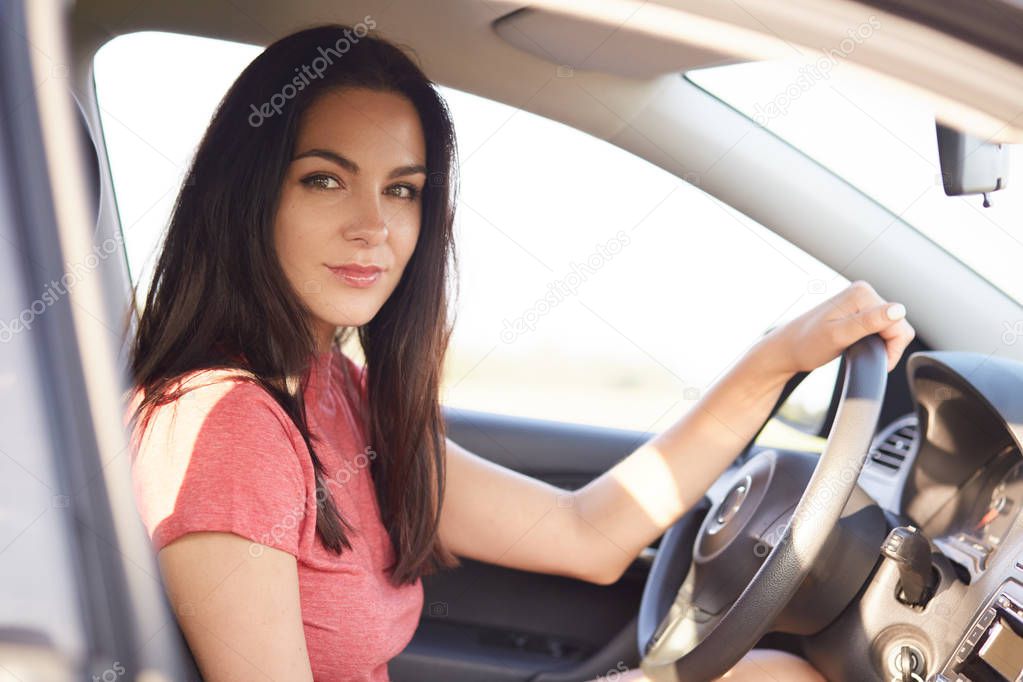 Sideways shot of good looking serious brunette girl drives car professionaly, sits on drivers seat, keeps hand on wheel, ready to cover long distance, dressed in casual clothing. Riding concept