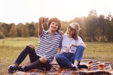 Açık shot memnun kız ve erkek arkadaşı birlikte sevinç var, ekose üzerinde oturup, akıllı telefon fotoğraf yapmak için poz, selfie sosyal ağlarda portre ve Paylaş görüntüler yapmak, doğa keyfini çıkarın