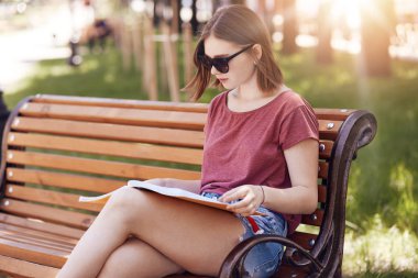 Ciddi öğrenci yatay görünüm trendy güneş gözlüğü, rahat t gömlek giyer ve jean şort, ders kitabı, konsantre oturur bankta, pozlar Park, öğrenme ve eğitim ile meşgul. Eğitim kavramı