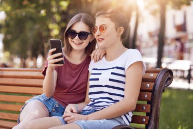 Sosyal ağlar, kullanım akıllı telefonu için eğlence, iletide mutlu genç kız fotoğrafı trendy güneş gözlüğü, poz tahta bankta açık, kablosuz internet'e bağlı giyer. Boş zaman kavramı