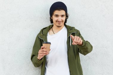 Yakışıklı olan memnun samimi ifade ile doğrudan kamera işaret parmağı ile gösterdiğine, moda ceket giymiş, himeseld sıcak içecek beyaz izole ile soğuk havalarda sıcak