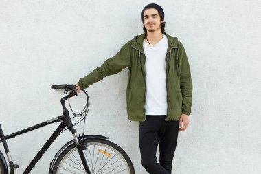 Aktif erkek bisikletçi el onun Bisiklet gidon üzerinde tutar, yeşil ceket, siyah şapka giyer, beyaz beton duvara teşkil etmektedir, boş zaman harcıyor, hobi seviyor. Sakallı şık adam mesafe kapsar