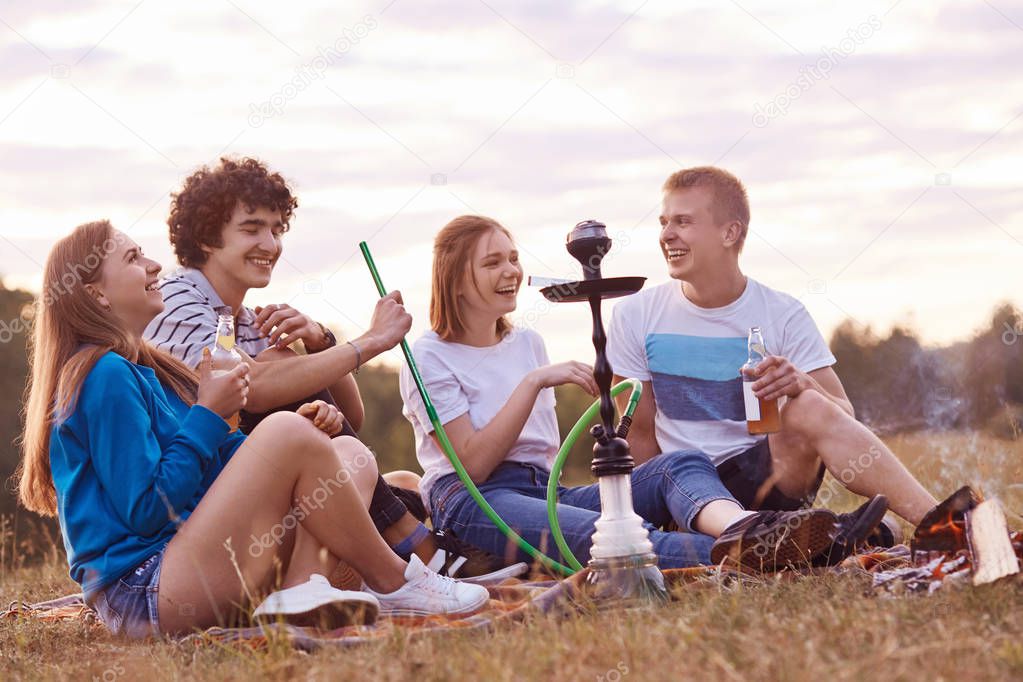 Two couple have joy together while have picnic outdoor, smoke hookah, drinks energetic beverages, being in high spirit, tell each other funny life stories. Outdoor shot. Relationship concept