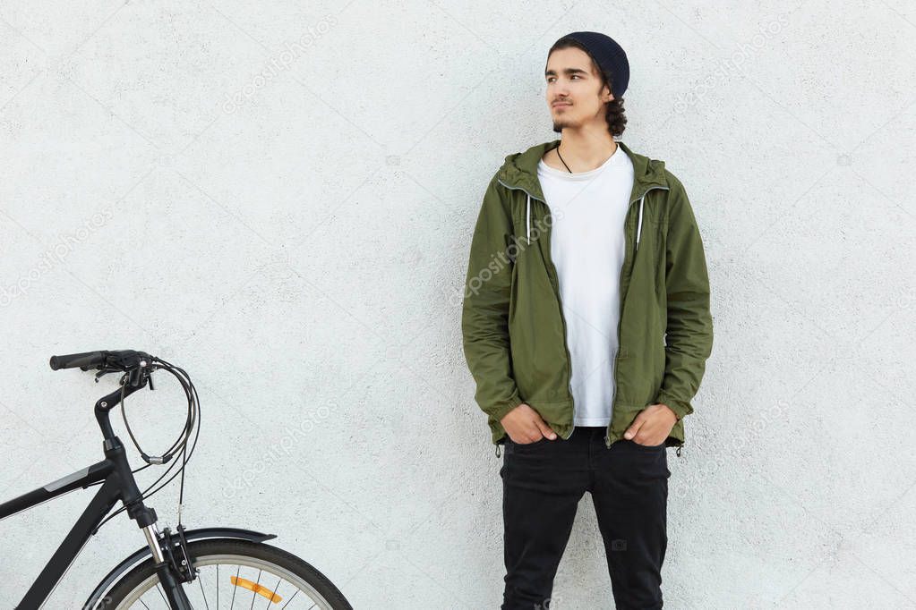 Horizontal shot of male cyclist dressed in fashionable clothes, holds hands in pockets, stands near his bicycle against white concrete wall, looks aside with thoughtful expression. Hobby concept