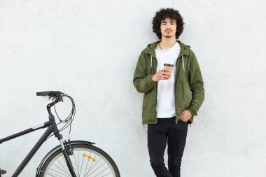 Giyinmiş moda ceket, siyah pantolon, içecekler sıcak içecek, kıvırcık saçlı yakışıklı biri kapalı atış Bisiklet beyaz beton duvara yakın duruyor. İnsanlar, Bisiklet ve yaşam tarzı kavramı