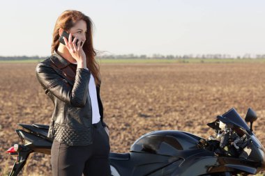 Kadın motorcu telefon görüşmesinde, durur yol, motosiklet yakınındaki pozlar, arkadaş, deri ceket ve siyah pantolon giymiş dicusses son haberleri var. Teknolojisi ve ulaşım kavramı