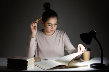Busy student absorbed in reading, wears round transparent glasses for good vision, uses reading lamp, sits at workplace, works in darkness, holds pencil, has clever look, prepares for final exam clipart