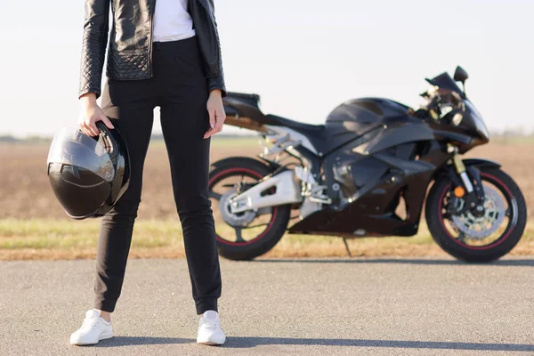 黒革のジャケットに身を包んだ女性バイカーの水平方向のショットをトリミング ズボン 白色スニーカー ヘルメットを保持して アスファルト バック グラウンドでクールな高速バイクの上に立ちます アウトドア ライフ スタイル — ストック写真