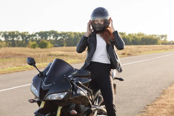 スタイリッシュな自信を持って女性バイカーの水平視野ファッショナブルな服を着て バイクに座って ヘルメットを身に着けている 長い先をカバー 屋外ポーズします モーターサイク リストは 屋外の旅行を楽しんでいます — ストック写真
