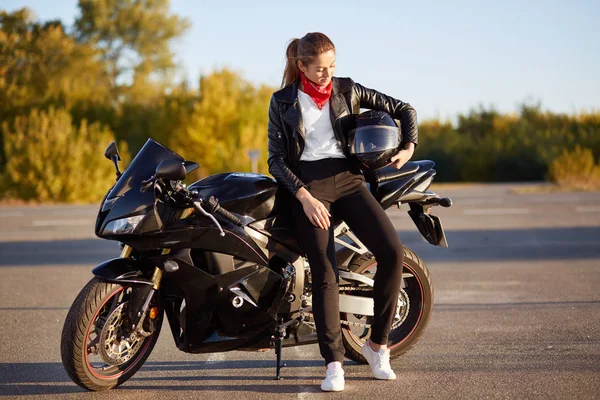 若い女性のモーターサイク リストの写真おしゃれな服 白いスニーカーを身に着けている 腕の下でヘルメットを保持して 黒バイクの近くに立って アスファルトの上に立つ 屋外ポーズします とライフ スタイルのコンセプト — ストック写真