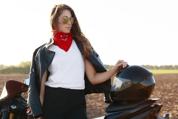 思慮深いブルネットの女性身に着けているスタイリッシュな革ジャケット トレンディな色合いの横のショットがバイクのそばに立つ ヘルメット 顔はともかく真面目に続けて手がアクティブなリゾート 人と極端な概念 — ストック写真