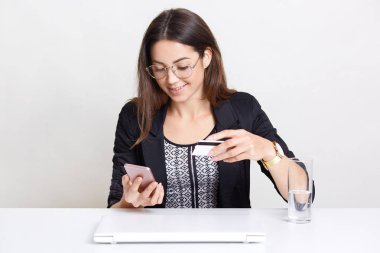 Neşeli kadın siyah giysilerle giyinmiş şeffaf gözlük takar, modern cep telefonu, plastik kart, maaş almak için mutlu tutar, beyaz arka planı teşkil etmektedir, suyu içer, uzaktan çalışır