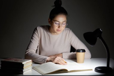 Meşgul kadın genç kitap okur, Masa lambası kullanır, son sınav için hazırlar, masaüstü oturur, görme duyusuyla ilgili yuvarlak gözlük takar, koyu arka planı teşkil etmektedir. Okuma akşamları. Eğitim kavramı